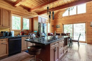 A wooden cabin kitchen with a large island, wooden cabinets, and a dining area with a view of the forest.