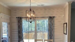 A dining area with a wooden table and chairs, a chandelier, and a view of the outdoors through a window with curtains. - 2027 Masters Rental Augusta GA (Harlem)