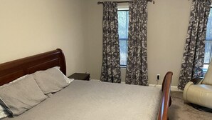 A bedroom with a wooden bed, a nightstand, a chair, and floral curtains. - 2027 Masters Rental Augusta GA (Harlem)