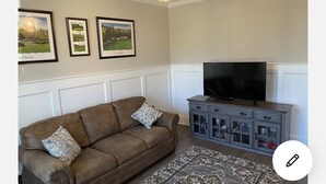 A living room with a brown sofa, patterned cushions, a flat-screen TV, and a gray cabinet with glass doors. - 2027 Masters Rental Augusta GA (Harlem)