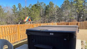 A hot tub with a wooden deck and a chair, surrounded by a wooden fence and trees. - 2027 Masters Rental Augusta GA (Harlem)