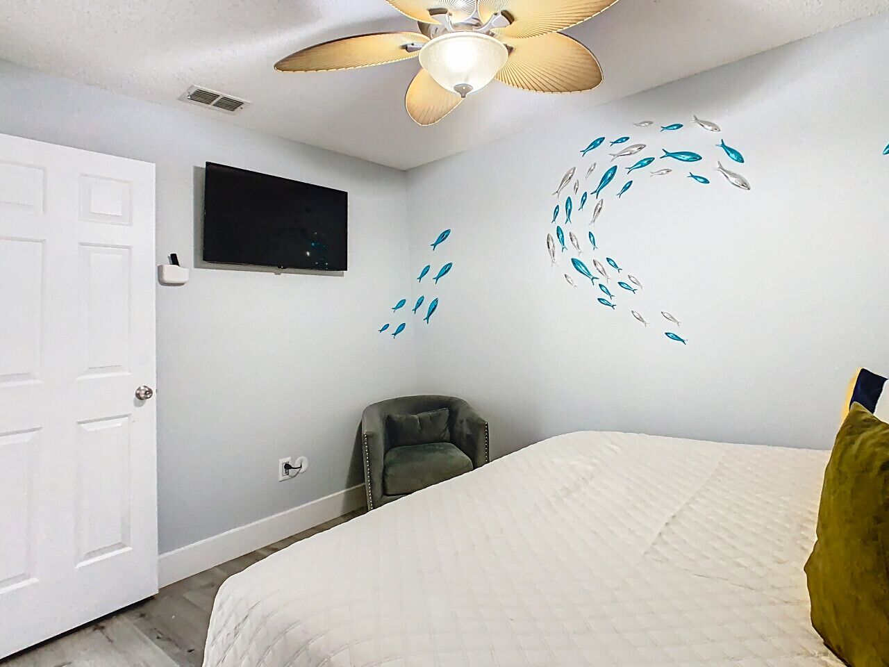 Un dormitorio con una cama, ventilador de techo, televisor montado en la pared y una silla.