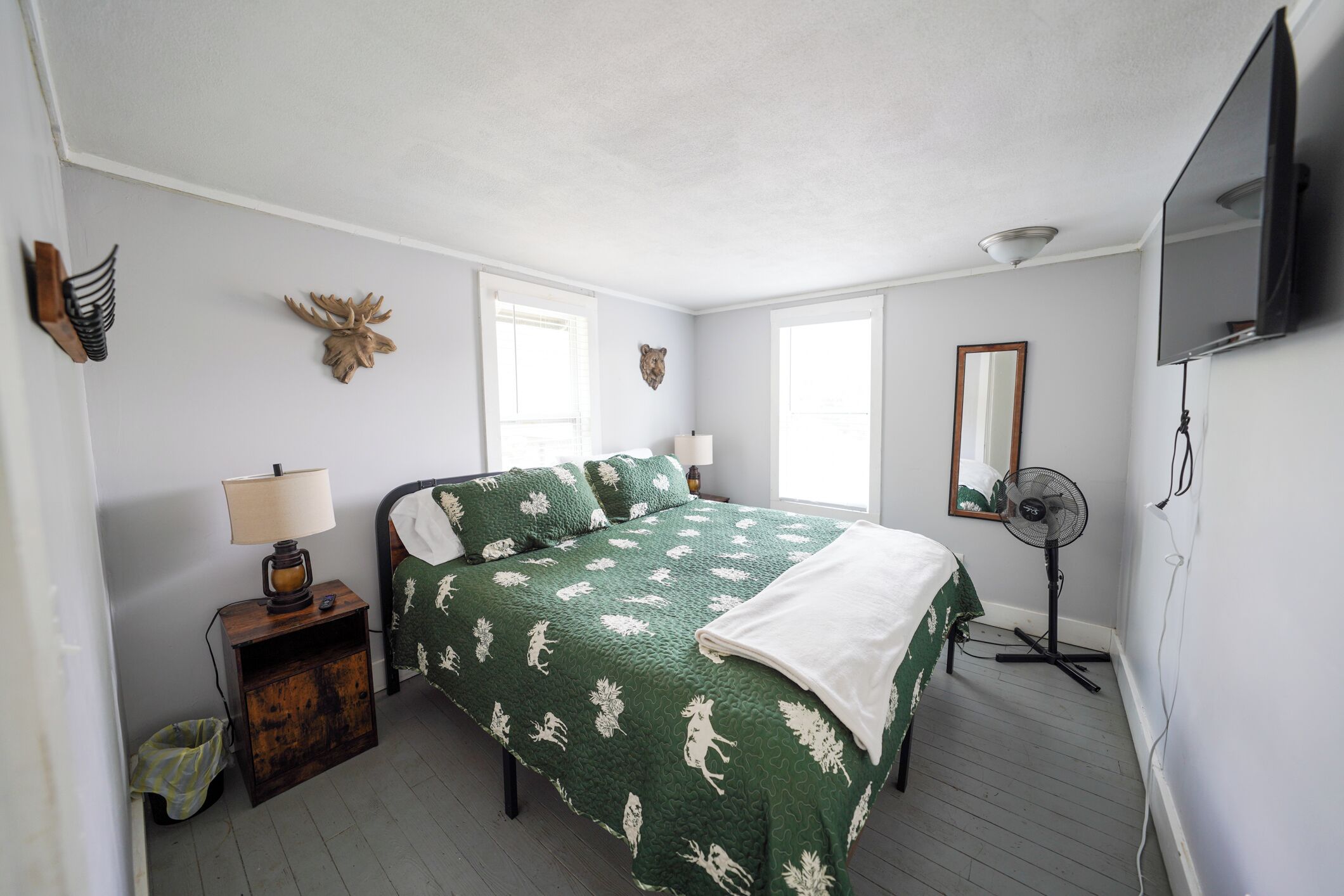 A bedroom with a green quilted bedspread, a bedside table with a lamp, a wooden nightstand, a small trash bin, a wall-mounted TV, and a standing fan.