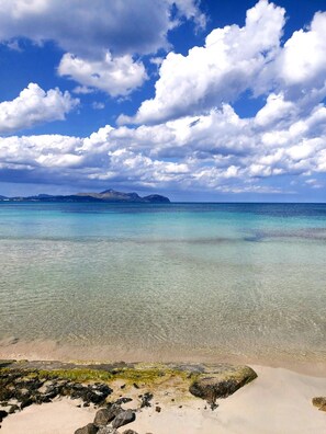 A beach with clear turquoise water, rocky shoreline, and distant mountains under a partly cloudy sky.