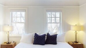 Un dormitorio con una cama, dos mesitas de noche, dos lámparas y dos ventanas.