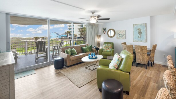 A spacious living room with a large sliding glass door leading to a balcony, a ceiling fan, and a dining area with wicker furniture. - Sandcastle Condominiums Unit 113 (Port Aransas)