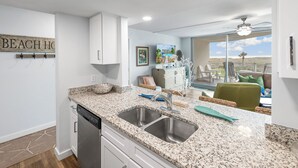 A kitchen with granite countertops, stainless steel sinks, and white cabinetry. There is a dining area with a view of the landscape. - Sandcastle Condominiums Unit 113 (Port Aransas)
