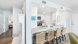 A modern kitchen with white cabinets, a granite countertop, and a built-in microwave. There are wicker chairs around the island and a green bench in the corner. - Sandcastle Condominiums Unit 113 (Port Aransas)
