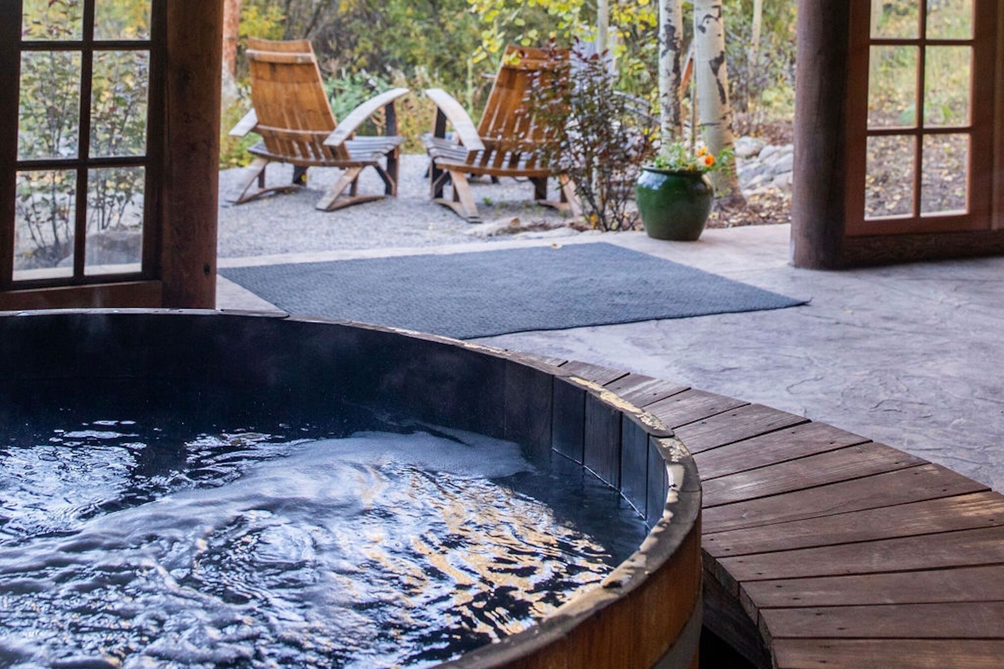 A hot tub with a wooden deck, two wooden chairs, and a potted plant in a covered outdoor area.