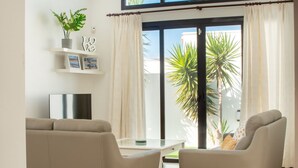 A modern living room with a sofa, armchair, and a glass coffee table. There is a TV on a stand, a plant on a shelf, and a sliding glass door leading to a balcony with a palm tree. - VILLA ORCADES CLOSE TO TOWN CENTER POOL & A/C (Playa Blanca)