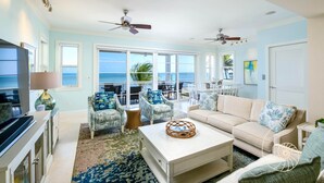 A spacious living room with a large window overlooking the ocean, a white sofa, patterned armchairs, and a television.