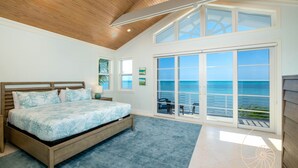 A bedroom with a large bed, a view of the ocean through a sliding glass door, and a wooden ceiling.