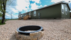 A modern building with a stone fire pit in the foreground, wooden deck, and a gravel driveway.