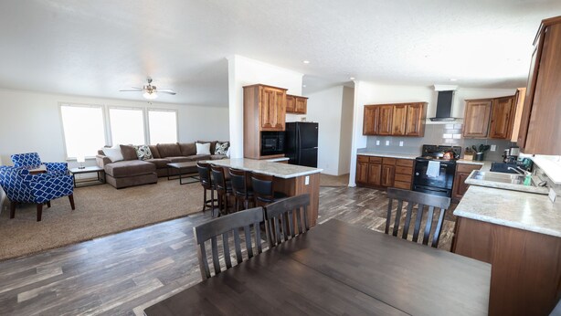 A spacious living room with a large sofa and a dining area with a wooden table and chairs. The kitchen features wooden cabinets and a central island with a sink.