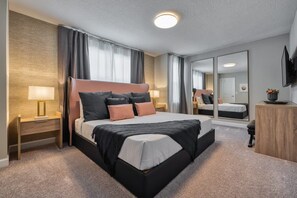 Une chambre d’hôtel moderne avec un grand lit, des tables de chevet avec des lampes, une tête de lit en bois, un miroir et un téléviseur posé sur une commode en bois.