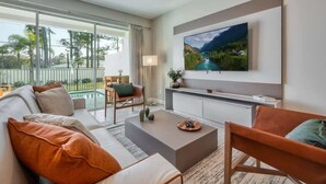 Una sala de estar moderna con un sofá, una mesa de centro y sillas. Un televisor de pantalla plana está montado en la pared, y una ventana ofrece vistas al exterior.