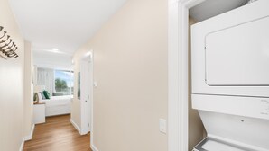 Un couloir doté d’un plancher en bois, d’un mur blanc et d’une porte blanche qui donne sur une pièce avec une fenêtre et une vue sur l’extérieur.