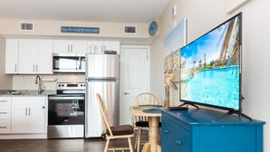 Une cuisine moderne avec des armoires blanches, des appareils en acier inoxydable, un coin repas avec une chaise et une table en bois, et un téléviseur diffusant une scène de plage.