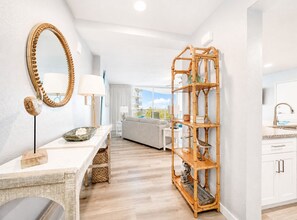 A modern hotel room with a large mirror, a wooden console table, a sofa, a bamboo shelving unit, and a kitchen area with white cabinets and a sink.