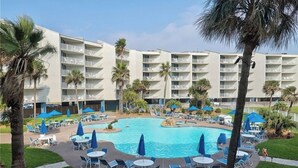 Et hotel med en central swimmingpool omgivet af liggestole og parasoller, palmer og en bygning med mange etager og altaner.