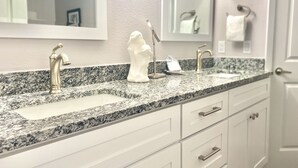 A bathroom with a granite countertop, two sinks, a towel rack, and a mirror. - Sandcastle Condominiums Unit 202 (Port Aransas)