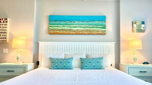 A hotel room with a large bed, two bedside tables with lamps, a wall-mounted painting of a beach, and a sign with text. - Sandcastle Condominiums Unit 202 (Port Aransas)