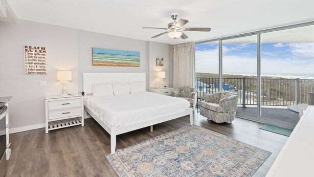 A bedroom with a large bed, a ceiling fan, a view of the ocean, a balcony with a chair, and a beach rules sign. - Sandcastle Condominiums Unit 202 (Port Aransas)