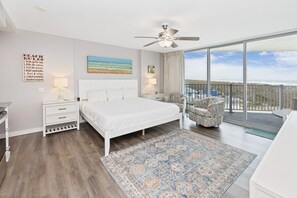 Un dormitorio con una cama grande, un ventilador de techo, vista al océano, un balcón con una silla y un cartel con las normas de la playa.