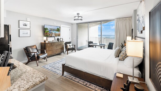 Een moderne hotelkamer met een groot bed, een bureau, een stoel, een dressoir en een televisie. Er is een balkon met uitzicht op de oceaan.