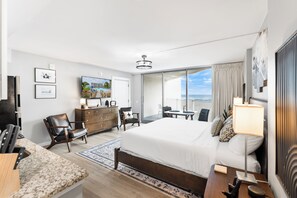 Une chambre d’hôtel moderne, dotée d’un grand lit, d’un bureau, d’une chaise, d’une commode et d’une télévision. Elle dispose également d’un balcon avec vue sur l’océan.