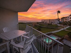 Un balcon offrant une vue sur l’océan au coucher du soleil, une piscine et un immeuble doté de nombreux balcons.