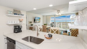 A modern kitchen with a large sink, white cabinets, and a coffee maker. There is a dining area with a table and chairs, and a balcony with a view of the ocean. - Sandcastle Condominiums Unit 213 (Port Aransas)