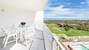 A balcony with a white table and chairs, a flower vase, and a view of a golf course and ocean. - Sandcastle Condominiums Unit 213 (Port Aransas)