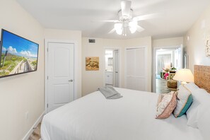 A bedroom with a large bed, a ceiling fan, a painting of a beach, and a view into the bathroom. - Sandcastle Condominiums Unit 213 (Port Aransas)