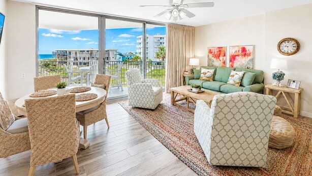 A spacious living room with a balcony overlooking a beach, a green sofa, and a dining area with wicker chairs. - Sandcastle Condominiums Unit 213 (Port Aransas)