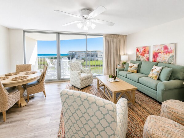 A spacious living room with a balcony, a green sofa, patterned chairs, and a ceiling fan. - Sandcastle Condominiums Unit 213 (Port Aransas)