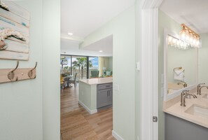 A modern kitchen with a large island, a dining area with a view, and a hallway with a wooden art piece and hooks. - Sandcastle Condominium Unit 218 (Port Aransas)