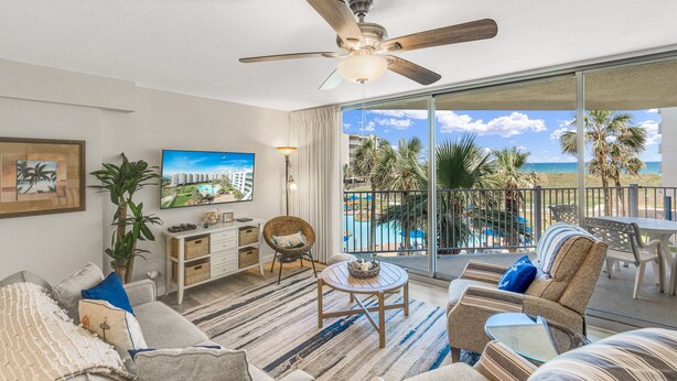 A spacious living room with a large TV, comfortable seating, a ceiling fan, and a balcony with a pool view. - Sandcastle Condominiums Unit 225 (Port Aransas)
