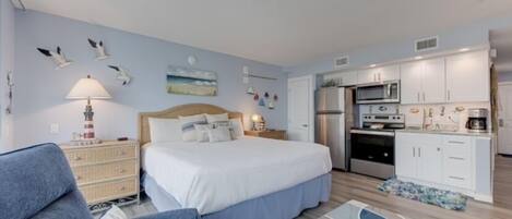 A bedroom with a bed, a nightstand, a lamp, and a dresser. There is a kitchenette with a microwave, oven, and refrigerator.