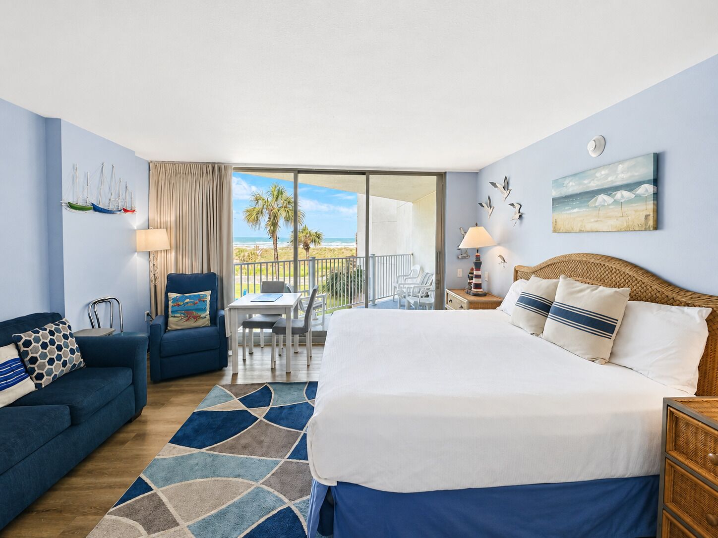 Habitación de hotel con cama, sofá, mesa de comedor, sillas, balcón con vista a la playa y un cuadro en la pared.