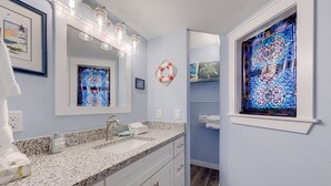 Une salle de bain dotée d’un comptoir en granit, d’armoires blanches, d’un grand miroir, d’un vitrail et d’un élément décoratif en forme de bouée de sauvetage.