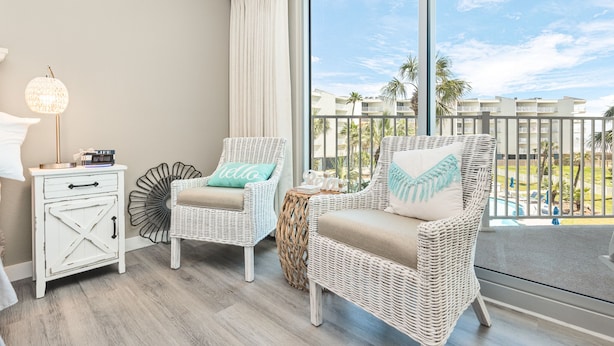 A balcony with wicker furniture, a white nightstand, and a view of a pool and buildings. - Sandcastle Condominiums Unit 227 (Port Aransas)