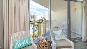 A balcony with wicker furniture, a small table with a book, and a view of a pool and buildings. - Sandcastle Condominiums Unit 227 (Port Aransas)