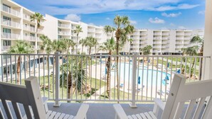 A balcony with white chairs overlooking a pool area with palm trees and a white building in the background. - Sandcastle Condominiums Unit 227 (Port Aransas)