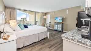 A hotel room with a large bed, a television, a desk with a computer, and a view of palm trees through the window. - Sandcastle Condominiums Unit 227 (Port Aransas)