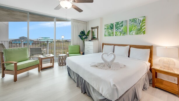 A hotel room with a large bed, a green sofa, a wooden nightstand, and a view of buildings through large windows.