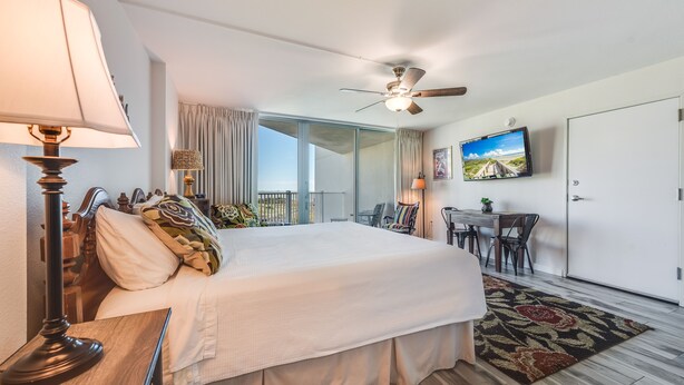 A hotel room with a large bed, a ceiling fan, a TV mounted on the wall, a dining table with chairs, and a view of the outdoors through a sliding door. - Sandcastle Condominiums Unit 235 (Port Aransas)