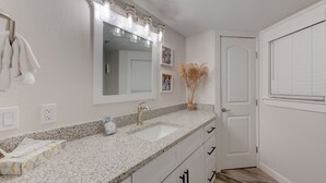 A bathroom with a granite countertop, a sink, a mirror with lights, a door, and a window with blinds. - Sandcastle Condominiums Unit 235 (Port Aransas)
