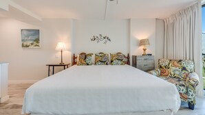 A bedroom with a large bed, two bedside tables with lamps, a floral-patterned armchair, and a painting on the wall. - Sandcastle Condominiums Unit 235 (Port Aransas)