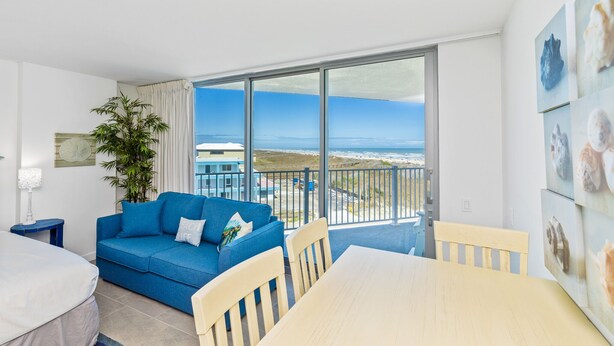 Ein Zimmer mit einem blauen Sofa, einem Holzesstisch und einem großen Fenster mit Blick auf den Strand und den Ozean.